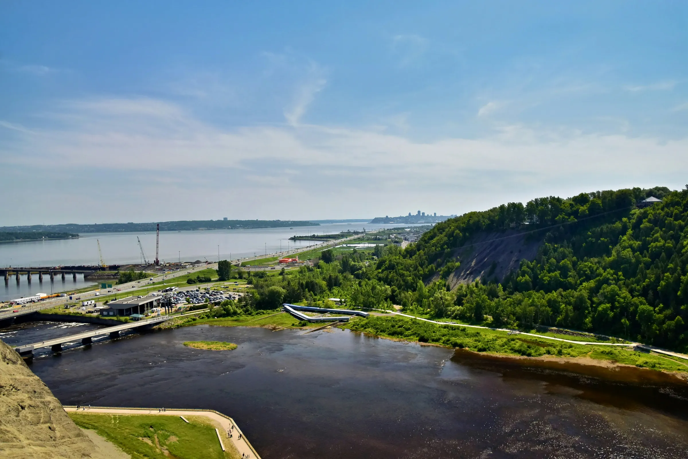 A wide river flowing past a forested cliff and a distant city skyline